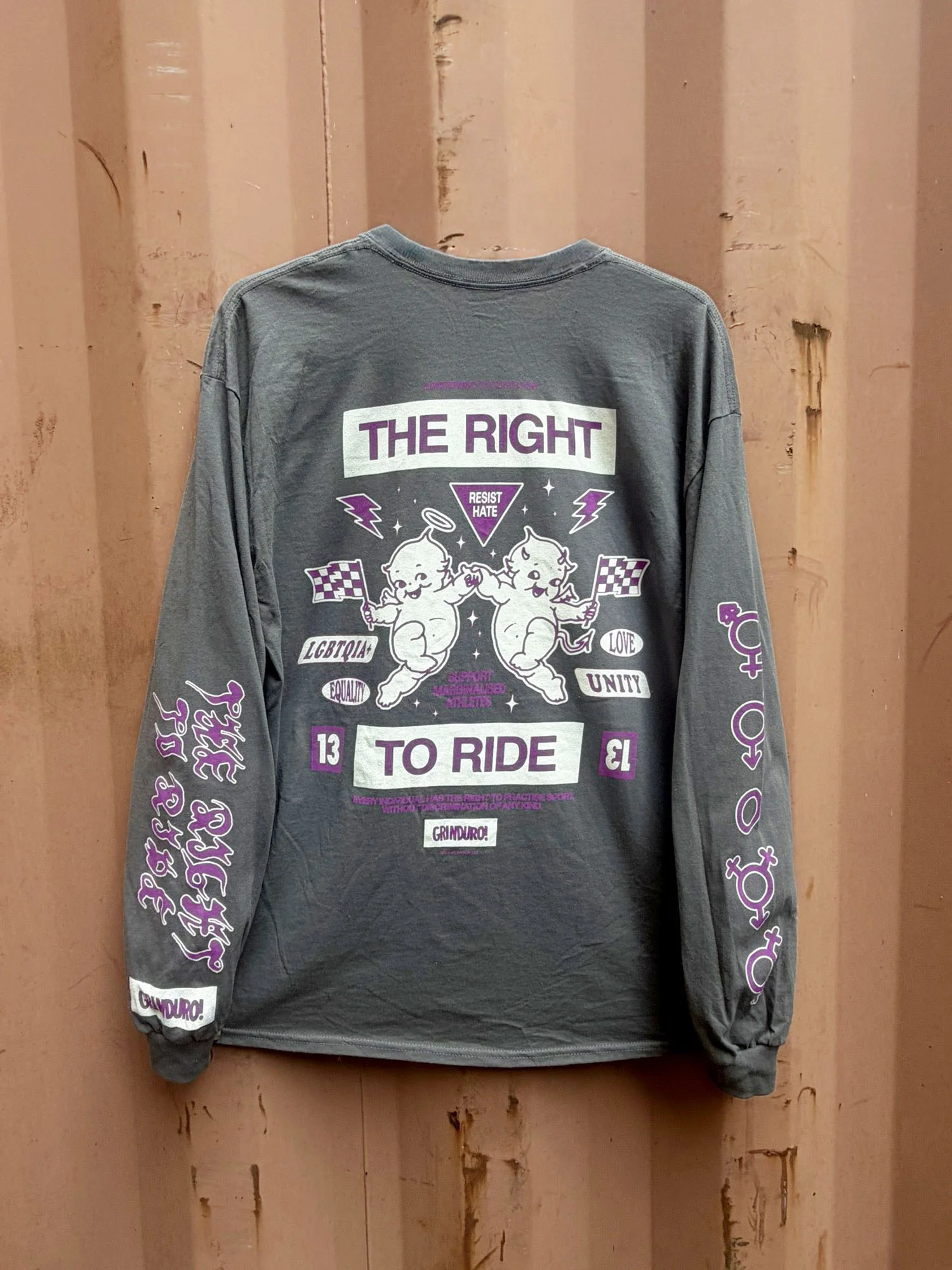 Back of a grey long-sleeve tee discharge printed with a purple and white 'The Right to Ride' design, hung against a rust-orange shipping container