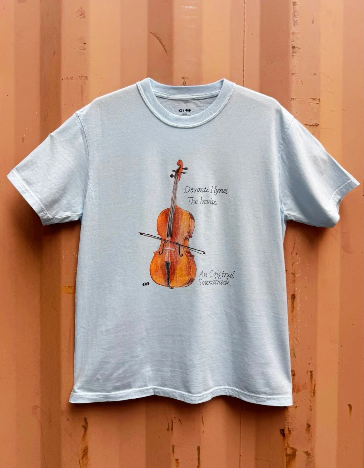 Pale blue enzyme-washed tee with a hand-drawn cello print for Devonté Hynes' The Invite, hung against a rust-orange shipping container